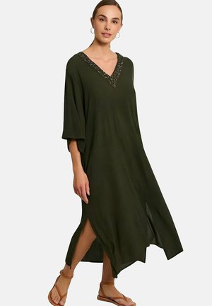 Femme portant une robe maxi ample vert foncé avec un décolleté en V orné de perles et des fentes latérales, associée à des sandales marron à bride cheville.