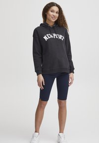 Felpa nera con la scritta "NEWPORT" in bianco ad arco, abbinata a pantaloni corti da bici blu navy. Il modello indossa sneakers bianche. Design semplice, vestibilità casual.