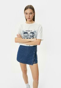 Camiseta gráfica blanca con la impresión "Brooklyn", combinada con una falda envolvente de mezclilla oscura. La modelo está de pie con los brazos cruzados, vistiendo calcetines blancos y zapatillas.