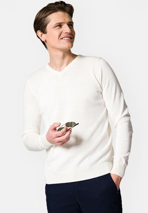 Męski biały sweter w serek z miękkiego materiału, z prążkowanymi mankietami i dołem, w połączeniu z ciemnymi spodniami i trzymanym w ręku okularami przeciwsłonecznymi.