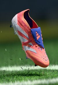 Botín de fútbol naranja y blanco con parte superior sintética, patrón texturizado y banda de Velcro azul. Presenta una distintiva marca Predator.