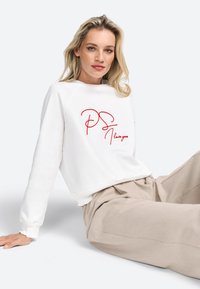Felpa bianca con testo ricamato rosso "PS I love you", con scollatura rotonda e vestibilità comoda. Modello che indossa pantaloni beige a gamba larga.