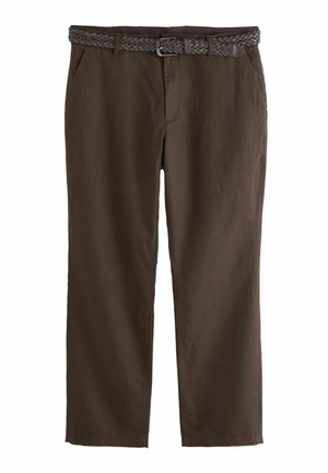 Mann mit Sonnenbrille, hellblau gestreiftem Hemd mit hochgekrempelten Ärmeln, brauner Hose und schwarzen Schuhen steht neben einer großen Topfpflanze im Innenbereich.