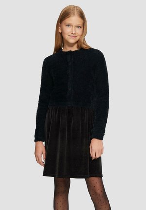 Zwarte fuzzy cardigan met een ronde hals, gecombineerd met een zwarte fluwelen jurk met een zachte textuur en subtiele glans, en gestippelde tights.