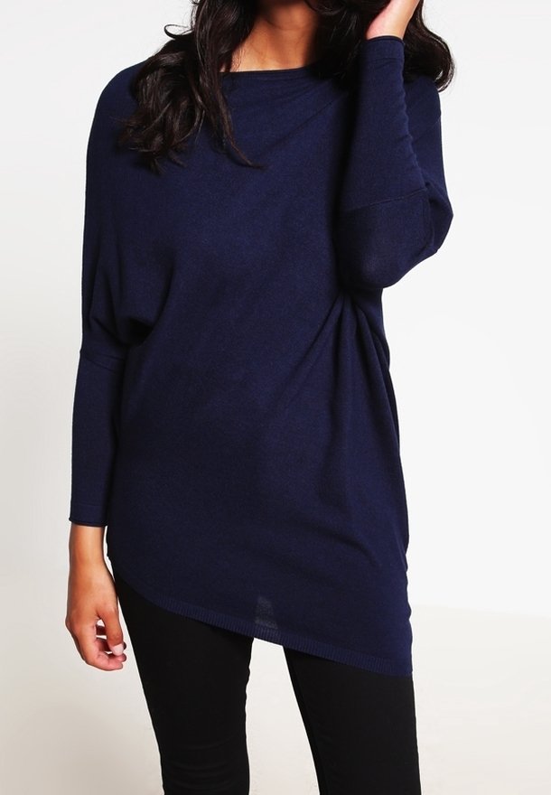 Marinblå oversized stickad topp med båthals och långa ärmar, med en draperad design och asymmetrisk fåll. Bärs med svarta byxor.