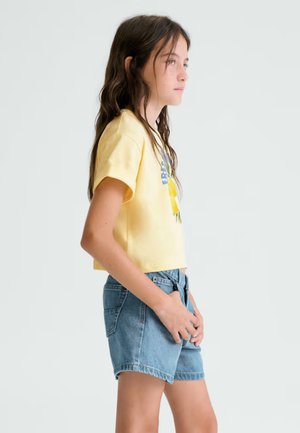 Ragazza con capelli castani lunghi che indossa una maglietta gialla a maniche corte e pantaloncini di jeans blu, in piedi di lato contro uno sfondo semplice.