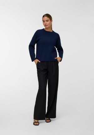 Pull-over en maille bleu marine avec un col rond, associé à un pantalon large noir. Le mannequin a les mains dans les poches et porte des sandales noires.