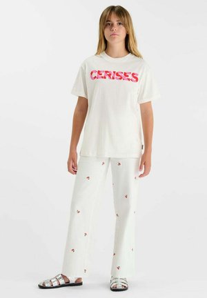 Jeune personne debout portant un pantalon blanc à petits motifs de cerises, une chemise blanche avec l'inscription "CERISES" en rouge, et des sandales argentées à bout ouvert.