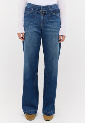 Person iført blå wide-leg jeans med et matchende denim bælte, en hvid langærmet skjorte og brune sko, stående mod en ensfarvet baggrund.