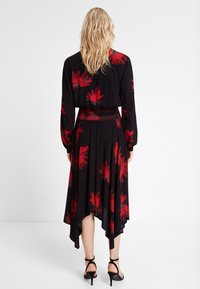 Musta mekko, jossa on punainen kukkakuvio, pitkät hihat, smokkivyötärö ja epäsymmetrinen helma, valmistettu kevyestä kankaasta.