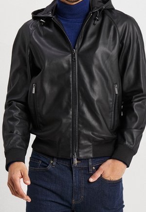 Man draagt een zwart leren jack met rits en capuchon over een blauwe coltrui en donkerblauwe spijkerbroek, handen dicht bij de zakken.