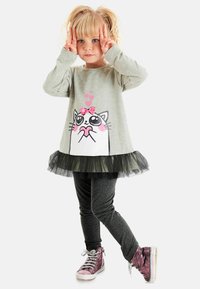 Grijze lange mouw top met een kat grafiek en een roze strik, gelaagd met een zwarte tule rok, gecombineerd met donkere leggings en glanzende roze sneakers.