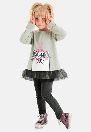 Grauer Langarmoberteil mit Katzenmotiv und rosa Schleife, kombiniert mit einem schwarzen Tüllrock, dazu dunkle Leggings und glänzende rosa Sneakers.