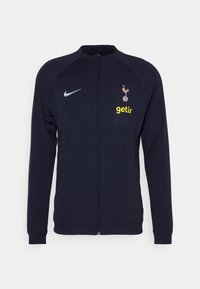 Giacca a track navy con chiusura a zip, logo Nike, emblema del Tottenham Hotspur e "getir" in giallo sul petto. Tessuto liscio e leggero.