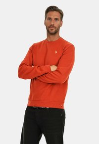 Oranges Sweatshirt aus weichem Material mit langen Ärmeln, gerippten Bündchen und einem kleinen gestickten Logo auf der Brust. Kombiniert mit schwarzen Hosen.