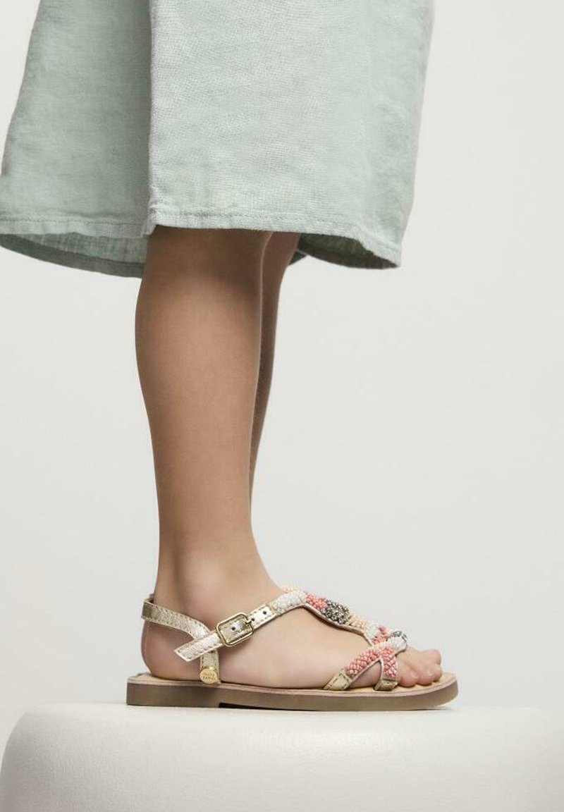 Sandalias con tiras y acabado metálico dorado, con acentos trenzados en rosa y blanco, y una hebilla plateada, sobre un fondo de tela clara.