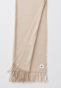 Écharpe beige clair en tissu doux, avec des bords à franges et une étiquette carrée avec un logo d'un côté.