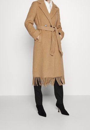 Femme portant un manteau ceinturé couleur camel avec des franges à l’ourlet, un pantalon noir et des bottines noires à talons, les mains dans les poches.