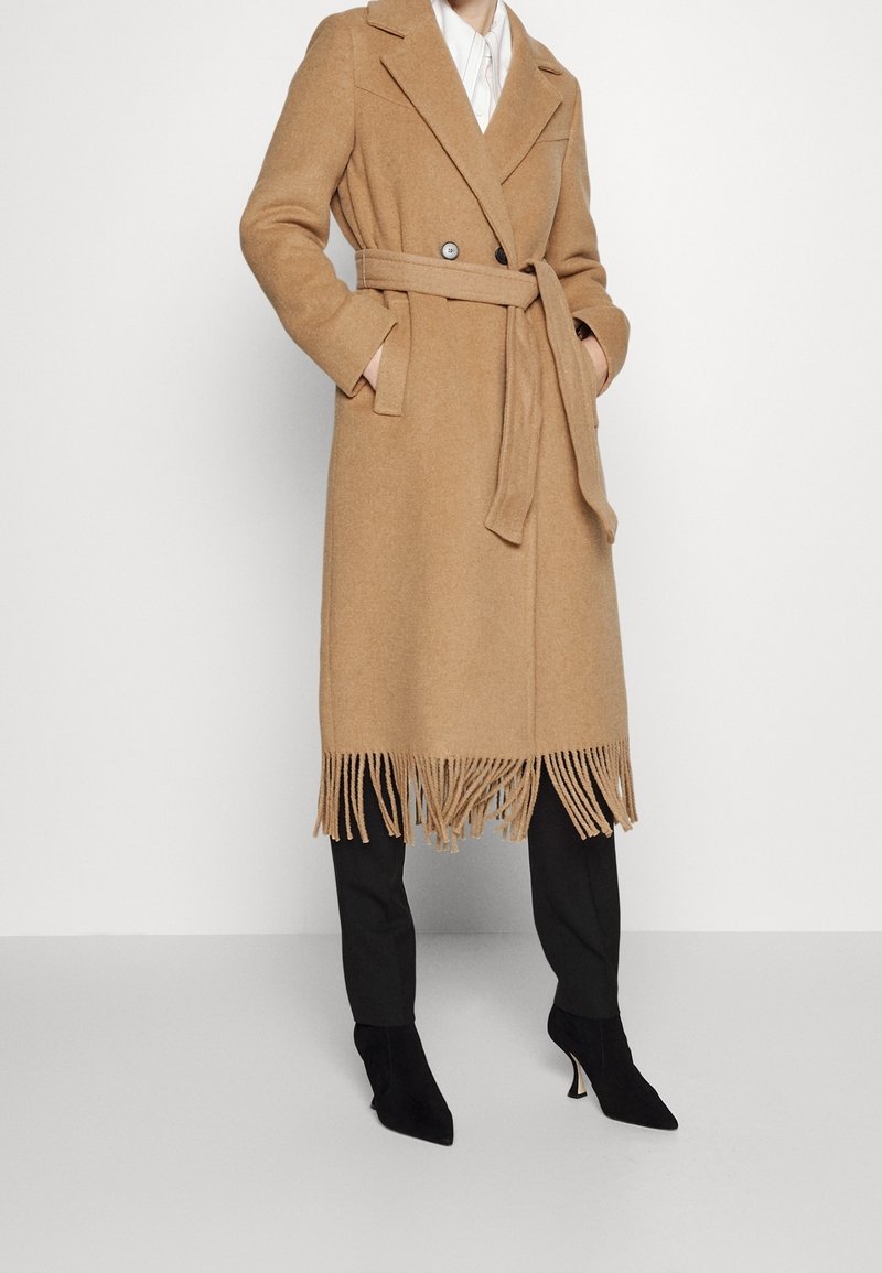 Femme portant un manteau ceinturé couleur camel avec des franges à l’ourlet, un pantalon noir et des bottines noires à talons, les mains dans les poches.