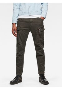 Mies, jolla on jalassaan tummanvihreät cargo-housut, joissa on useita taskuja, mustat nauhakengät, valkoinen paita ja vaalea farkkutakki, kädet taskuissa.