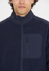 Marinblå fleecejacka med dragkedjeficka, med ett slätt tygpanel på den övre delen och en texturerad nedre sektion. Hög krage.
