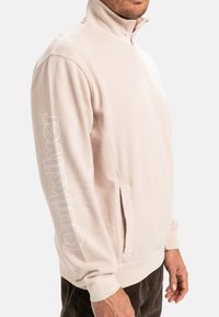 Light pink quarter-zip sweatshirt with a stand-up collar, featuring a logo on the left sleeve and front. Soft material and side pockets.