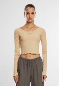 Beige gerippter langärmeliger Cropped-Top mit gewelltem Saum und KANI-Logo auf der Brust, kombiniert mit gerafften grauen Hosen.