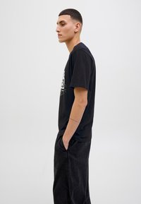 Sort bomulds T-shirt med korte ærmer og et grafisk print. Kombineret med mørke denimbukser med sidelommer og kontrastsyninger.