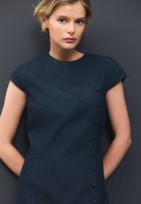 Robe en chambray bleu foncé à manches courtes, col rond, avec un détail de boutons sur le côté gauche. Texture lisse et coupe légèrement ajustée.