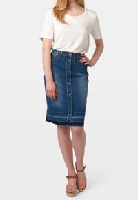 Falda lápiz de mezclilla con frente de botones, dobladillo sin terminar y costuras en contraste. Combinada con una blusa blanca y sandalias de tacón en color marrón.