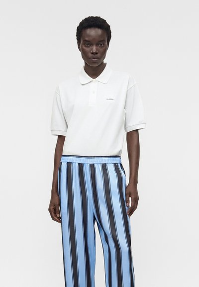 Άτομο που φορά άσπρο κοντομάνικο πόλο και μπλε παντελόνι με μαύρες και άσπρες κάθετες ρίγες, στέκεται μπροστά σε μονόχρωμο φόντο.