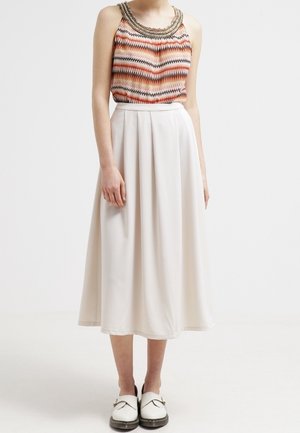 Kvinde bærer ærmeløs top med zigzag-mønster og perlebesat halsudskæring, cremefarvet høj-taljet plissert midi-nederdel og hvide loafers.
