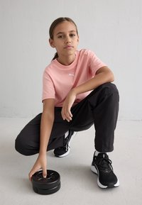 Ροζ βαμβακερό μπλουζάκι με λευκό λογότυπο Puma, μαύρο αθλητικό παντελόνι και μαύρα αθλητικά παπούτσια με λευκές λεπτομέρειες; χέρι που κρατάει μια μαύρη βά重量.