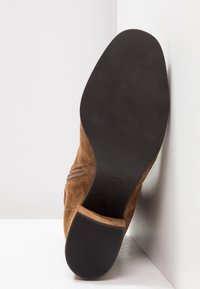 Botte en daim marron, positionnée en angle, montrant la semelle noire lisse et le talon en caoutchouc sur fond blanc.