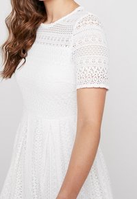 Biała sukienka z teksturowanej koronki, z krótkimi rękawami, z dopasowanym gorsem i zwiewną spódnicą, ozdobiona geometrycznymi wzorami.