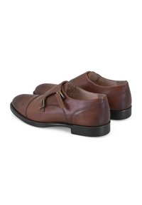 Bruine leren schoenen met dubbele gespsluiting, ronde neus, brogue-details en een zwarte rubberen zool. Gladde textuur met gestikte accenten.