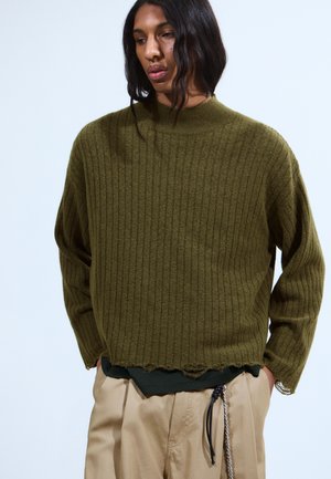 Personne avec de longs cheveux noirs portant un pull côtelé oversize olive et un pantalon beige, les mains dans les poches, devant un fond uni.