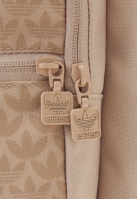Detail na béžovém batohu se zipem, vybavený dvěma béžovými plastovými táhly s vypouklým logem Adidas, texturovaná tkanina s jemným listovým vzorem.
