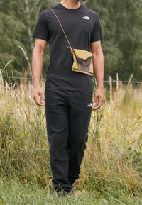 Man wearing black The North Face t-shirt and pants with a yellow crossbody pouch, standing in a grassy outdoor area near a wire fence.