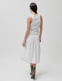 Γυναίκα με βρεγμένα μαλλιά, που φορά λευκή αμάνικη μπλούζα, λευκή φούστα μέσης μήκους και καφέ μποτάκια, περπατώντας μακριά πάνω σε ένα απλό φωτεινό φόντο.