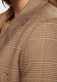 Brązowa marynarka w kratę z klapowanym kołnierzem, łącząca duże i małe wzory w kratę oraz teksturowane wykończenie tkaniny.