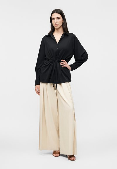 Γυναίκα με μακριά σκούρα μαλλιά φοράει μαύρο πουκάμισο με κουμπιά και ζώνη στη μέση, μπεζ παντελόνι με φαρδιά μπατζάκια και καφέ ψηλοτάκουνα πέδιλα με ανοιχτά δάχτυλα, στέκεται σε λευκό φόντο.