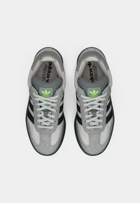 Ζευγάρι γκρι αθλητικά παπούτσια Adidas με μαύρες ρίγες, λευκά κορδόνια και φωσφορίζον πράσινο λογότυπο Adidas στη γλώσσα, σε άποψη από ψηλά σε λευκό φόντο.