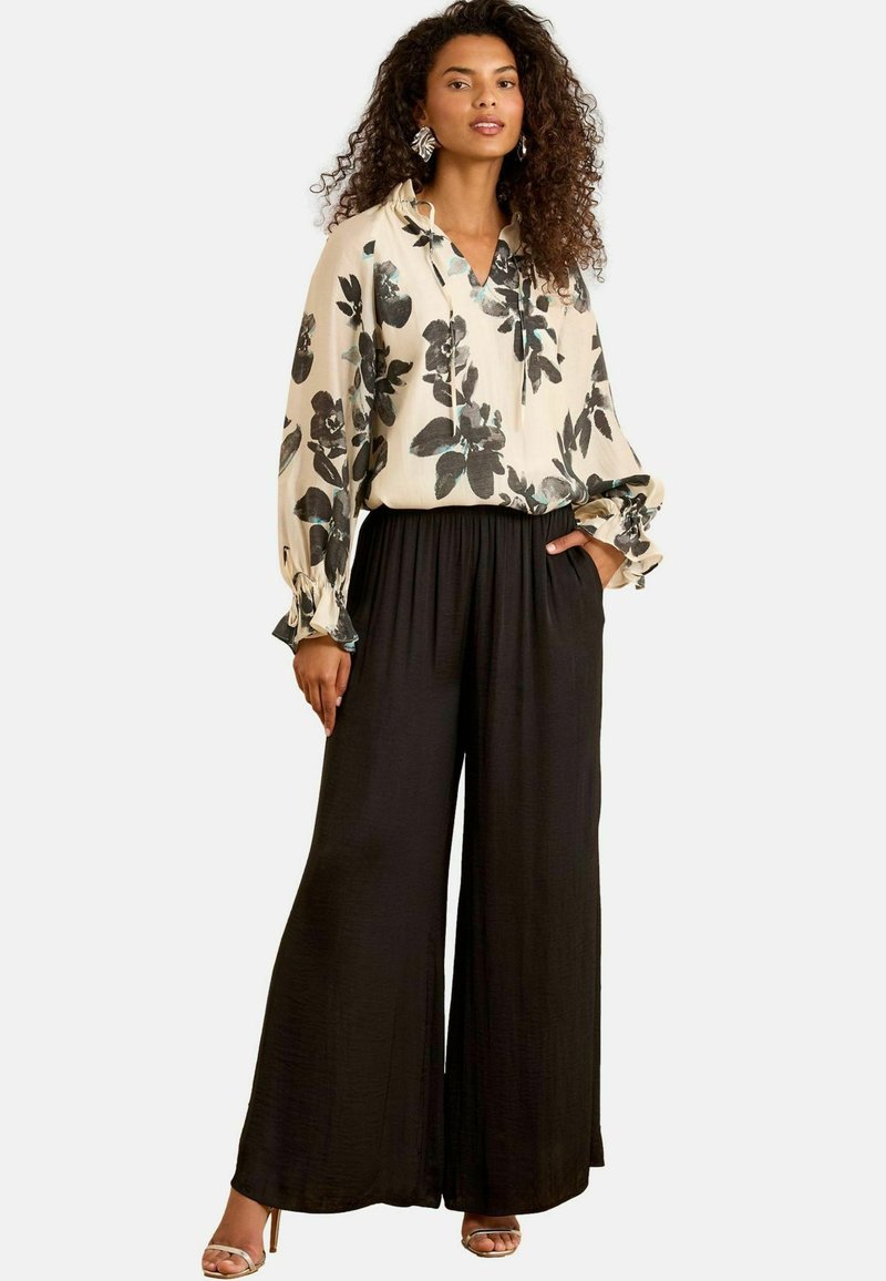 Blusa floreale color crema con fiori neri e verde teal, abbinata a pantaloni neri a gamba larga. Presenta una vestibilità comoda e una vita elastica.