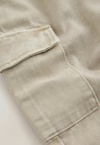 Gai beige audums ar ribota tekstūra, ar pārklājošu atloku kreisajā pusē, uz kura ir dūriena detaļa. Gluda, grumbu neizturīga virsma.