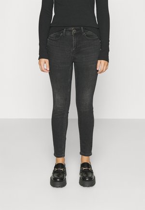 Jean slim en denim noir porté avec des mocassins noirs épais et un haut noir à manches longues, montré du milieu du torse jusqu’aux pieds.