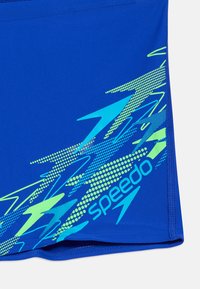 Μπλε ύφασμα μαγιό με αφηρημένο σχέδιο αστραπής σε νέον πράσινο και γαλάζιο και λογότυπο "speedo" σε γαλάζιο χρώμα.