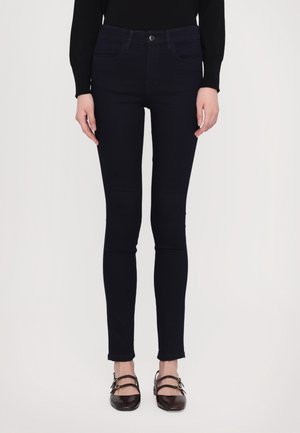 Mørkeblå skinny jeans med høj talje, to frontlommer og knaplukning. Bæres med brune sandaler med remme. Glat, strækbart stof.