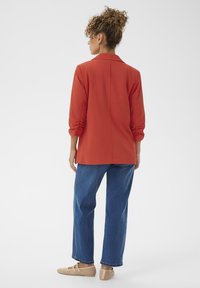 Chemise rouge à manches longues avec col, coupe décontractée et détail de couture au dos, associée à un jean droit bleu et des chaussures en toile beiges.