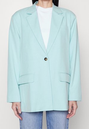 Person trägt ein hellblaues einreihiges Sakko mit einem Knopf über einem weißen Oberteil und blauen Jeans, steht mit entspannt an den Seiten hängenden Armen.